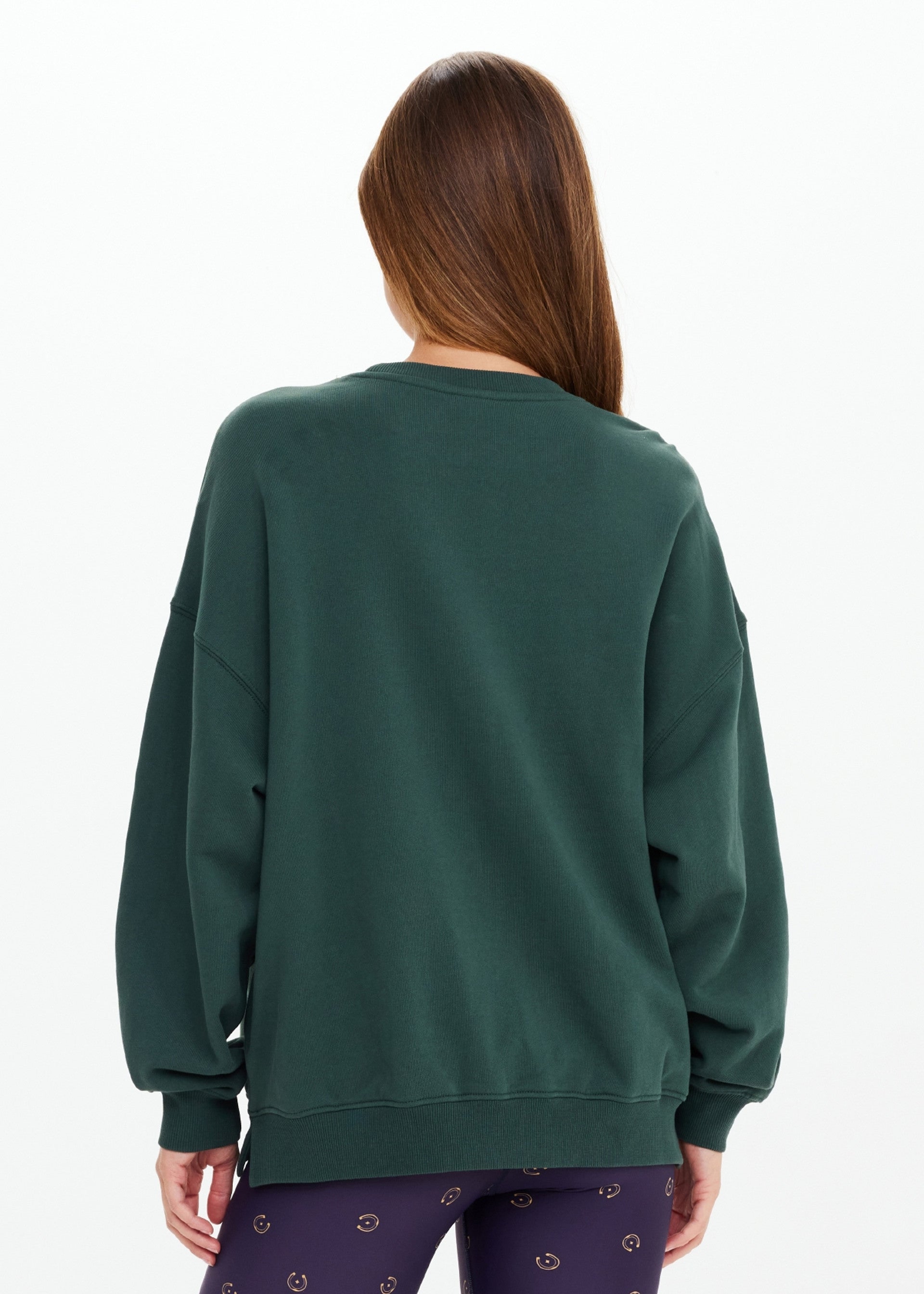 A person with long brown hair faces away, wearing The Upside's ALUMNI COOLUM CREW sweatshirt in dark green and patterned dark leggings, standing against a plain white background.