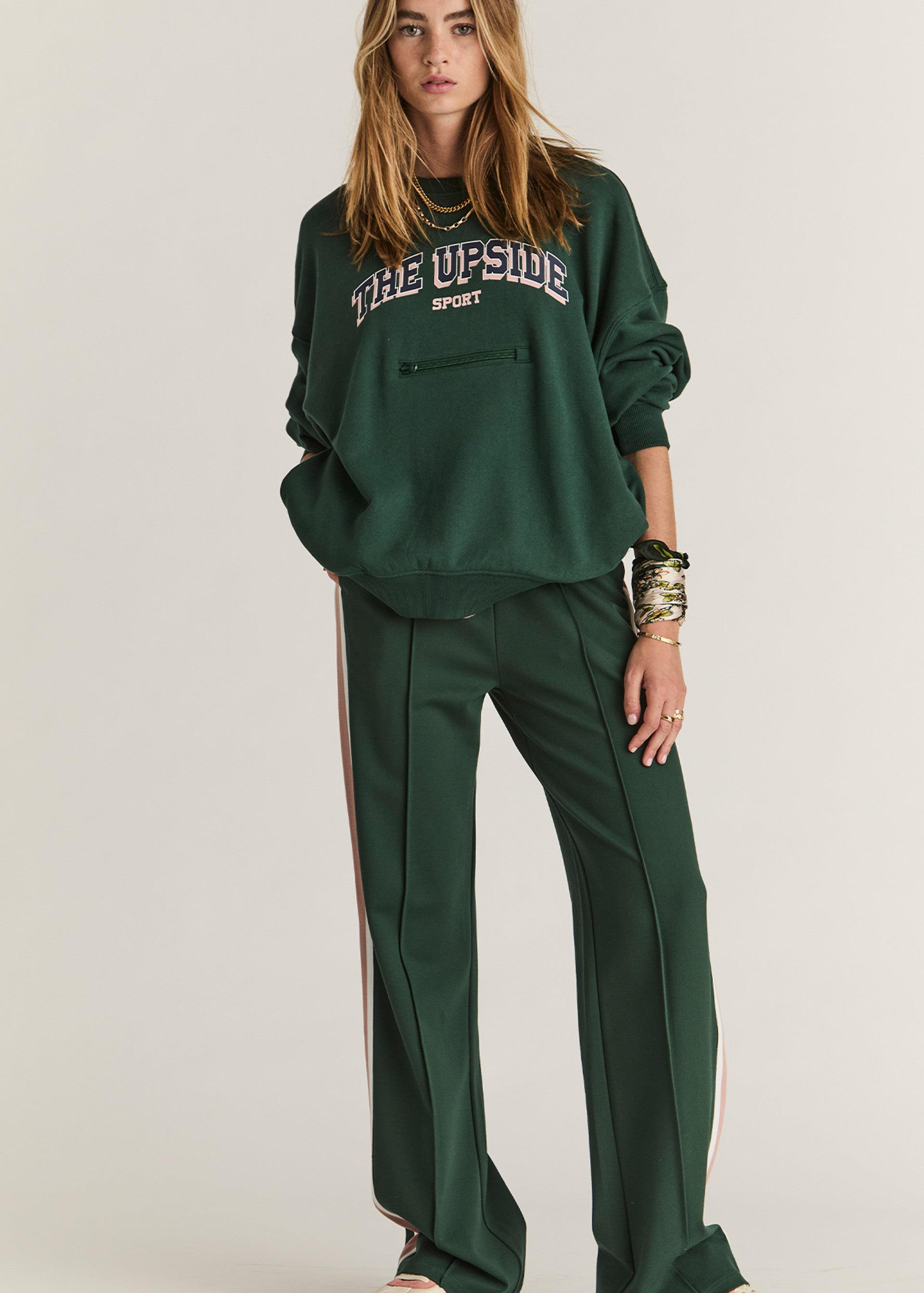 A woman sports The Upside's ALUMNI COOLUM CREW, a loose dark green organic cotton sweatshirt with "THE UPSIDE SPORT" print, paired with matching green wide-leg pants featuring white side stripes and gold bracelets, evoking vintage collegiate style.