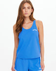 A woman with long brown hair wears The Upside’s WRANGLER ISSY TANK TOP and matching blue athletic shorts with white trim, standing against a plain white background.