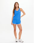 A woman poses in the WRANGLER ISSY TANK TOP by The Upside, styled with matching blue shorts and white running shoes against a plain white background.