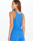 A woman with straight brown hair stands facing away, wearing The Upside's WRANGLER ISSY TANK TOP and matching shorts in blue performance mesh, set against a plain white background.