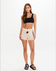 A woman with long, straight brown hair and a neutral expression wears The Upside's LUMINARY CROCHET HALI SHORT with an elasticated waist and a black crop top, styled with black sandals against a plain white background—perfect for relaxed beach days.