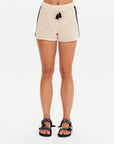 Wearing The Upside's LUMINARY CROCHET HALI SHORT—cream cotton crochet shorts with black side details and an elastic waist—paired with black patterned sandals, this relaxed look shines against a white background for the perfect beach day.