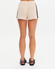 A woman faces away, wearing The Upside LUMINARY CROCHET HALI SHORT—cream cotton crochet shorts with an elastic waist and black trim—paired with black bow open-toe sandals, set against a plain white background.