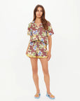 A woman stands against a plain white background, wearing The Upside's ISLA KLIO SHORT and matching short-sleeve button-up shirt with a brown base and bold yellow-blue floral print, paired with blue sandals.
