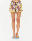 A person wearing The Upside ISLA KLIO SHORT with a brown floral pattern and yellow trim, paired with blue sandals featuring yellow straps, stands against a plain white background.