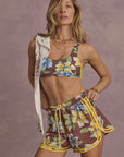 A woman models The Upside’s ISLA KLIO SHORT—a matching floral spin short with yellow trim—and a sports bra, draping a light shirt over her shoulder against a purple-gray textured backdrop.
