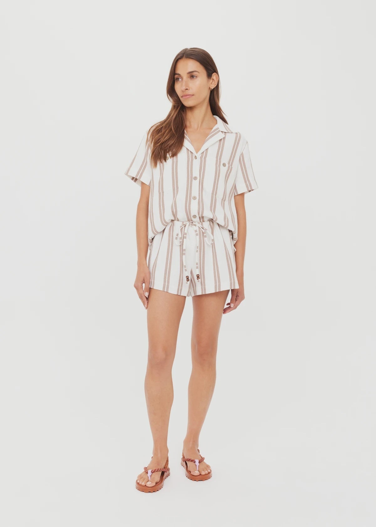 A woman stands against a white background wearing The Upside's BAJA ELIAS SHORT—a short-sleeved, button-up white romper with stripes and a tied waist—paired with brown sandals.