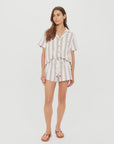A woman stands against a white background wearing The Upside's BAJA ELIAS SHORT—a short-sleeved, button-up white romper with stripes and a tied waist—paired with brown sandals.