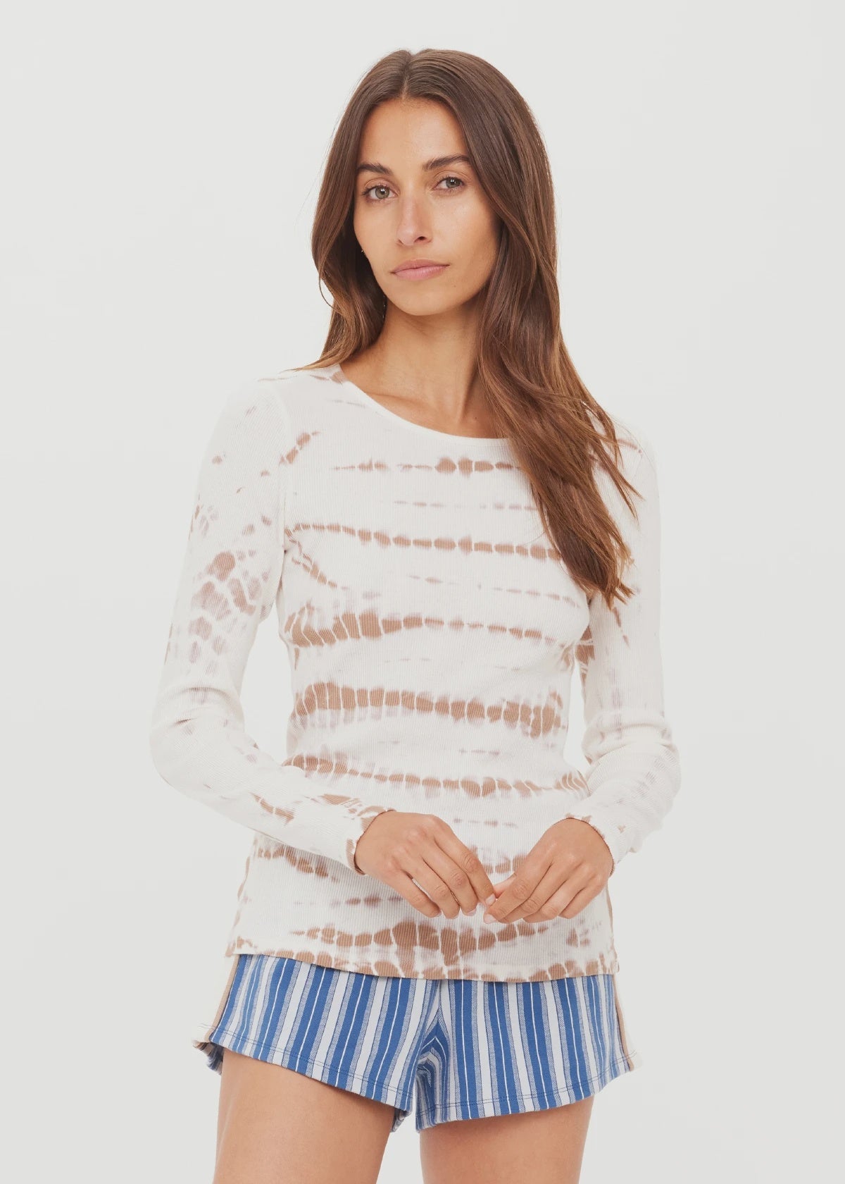 A woman with long brown hair wears The Upside’s AYLA CHRISSY LONG SLEEVE TOP and blue-and-white striped shorts, standing against a plain white background.