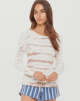 A woman with long brown hair wears The Upside’s AYLA CHRISSY LONG SLEEVE TOP and blue-and-white striped shorts, standing against a plain white background.