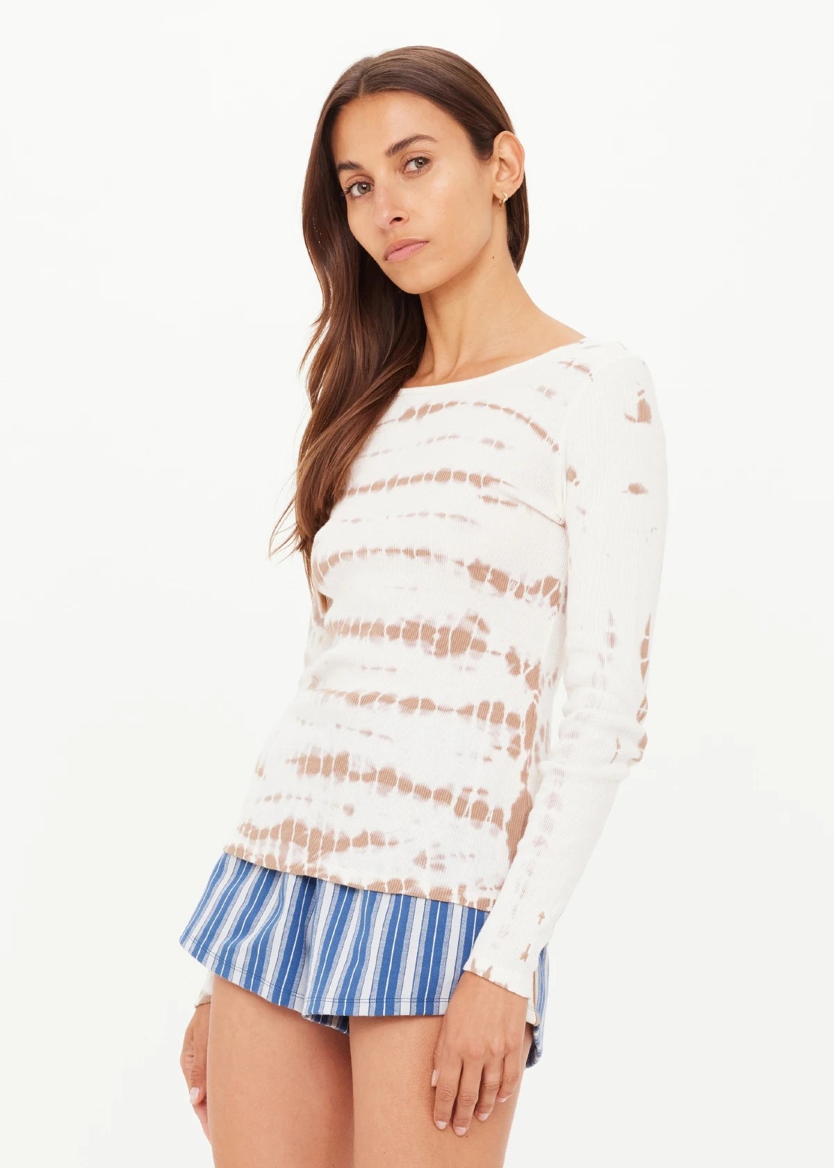 A woman with long brown hair wears The Upside AYLA CHRISSY LONG SLEEVE TOP, styled with blue-and-white striped shorts, standing against a plain white background.