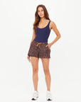 A woman with long brown hair stands against a white background wearing The Upside LUDLOW BILLIE RUN SHORT, featuring a leopard print and drawstring, paired with a navy tank top and white sneakers. One hand is in her pocket.