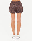 A person is shown from the waist down, wearing The Upside LUDLOW BILLIE RUN SHORT in leopard print recycled polyester with white sneakers, standing against a plain white background.