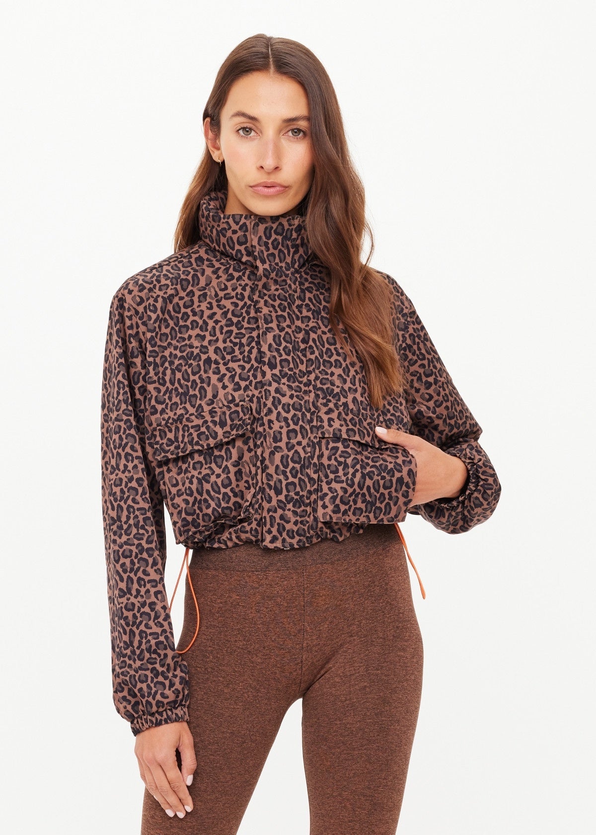 A woman with long brown hair models The Upside’s LUDLOW ACE SHELL JACKET, a cropped leopard print style with a high collar, paired with brown leggings against a plain white background.