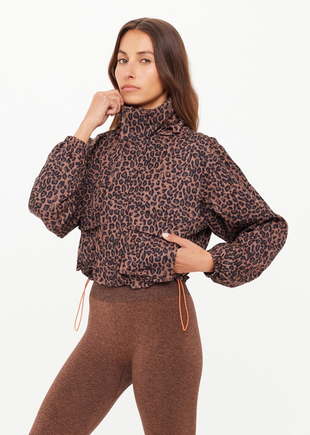 A woman wears The Upside LUDLOW ACE SHELL JACKET, featuring a brown leopard print and high collar, paired with brown leggings. She stands against a plain white background, looking at the camera with a neutral expression.