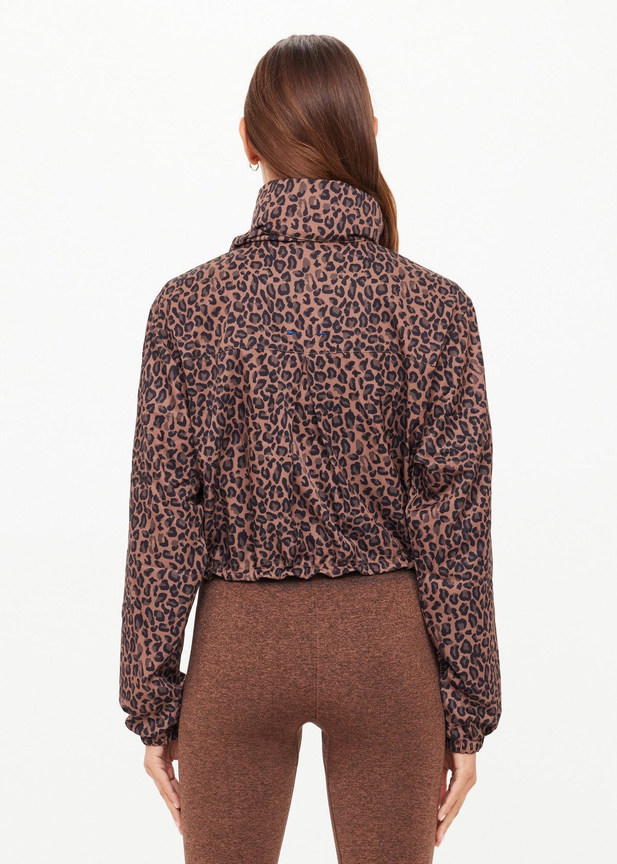 A woman with long brown hair is seen from behind, wearing The Upside's LUDLOW ACE SHELL JACKET in leopard print and brown leggings, standing against a plain white background.