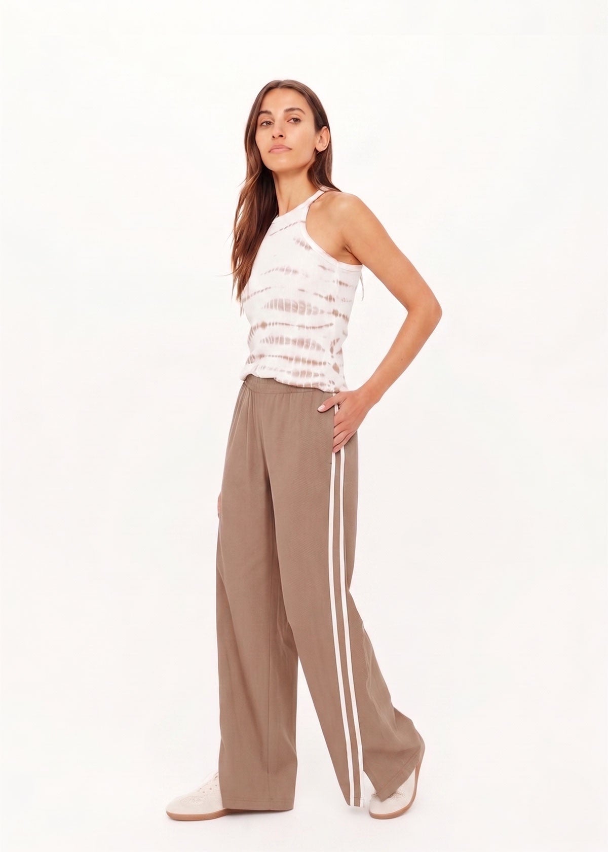 A woman with long brown hair poses confidently against a white backdrop, wearing The Upside’s JULIET PANT S26 with white side stripes, a sleeveless tie-dye tank top, and white sneakers. One hand rests on her hip.