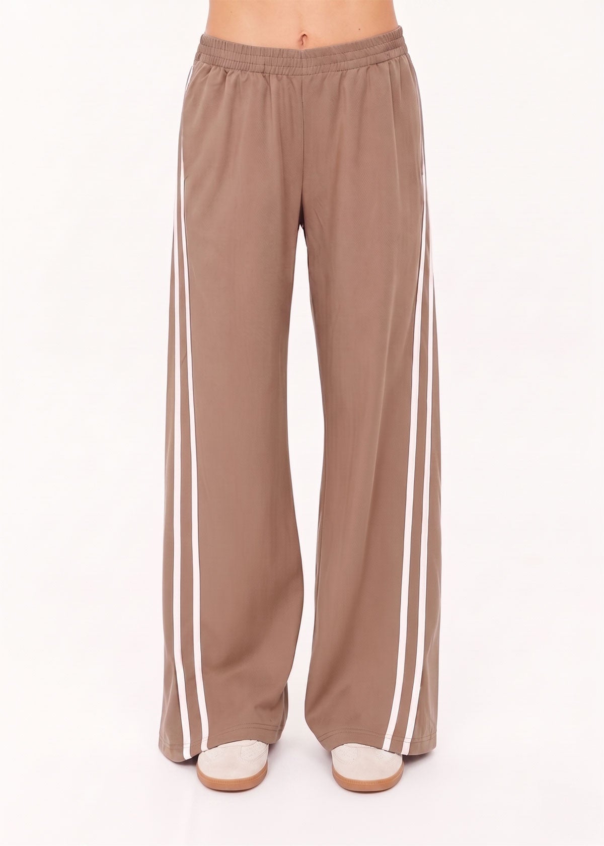A person wears The Upside JULIET PANT S26—loose, light brown twill pants with two vertical white side stripes, elasticated waist, and wide legs—standing against a plain white background.