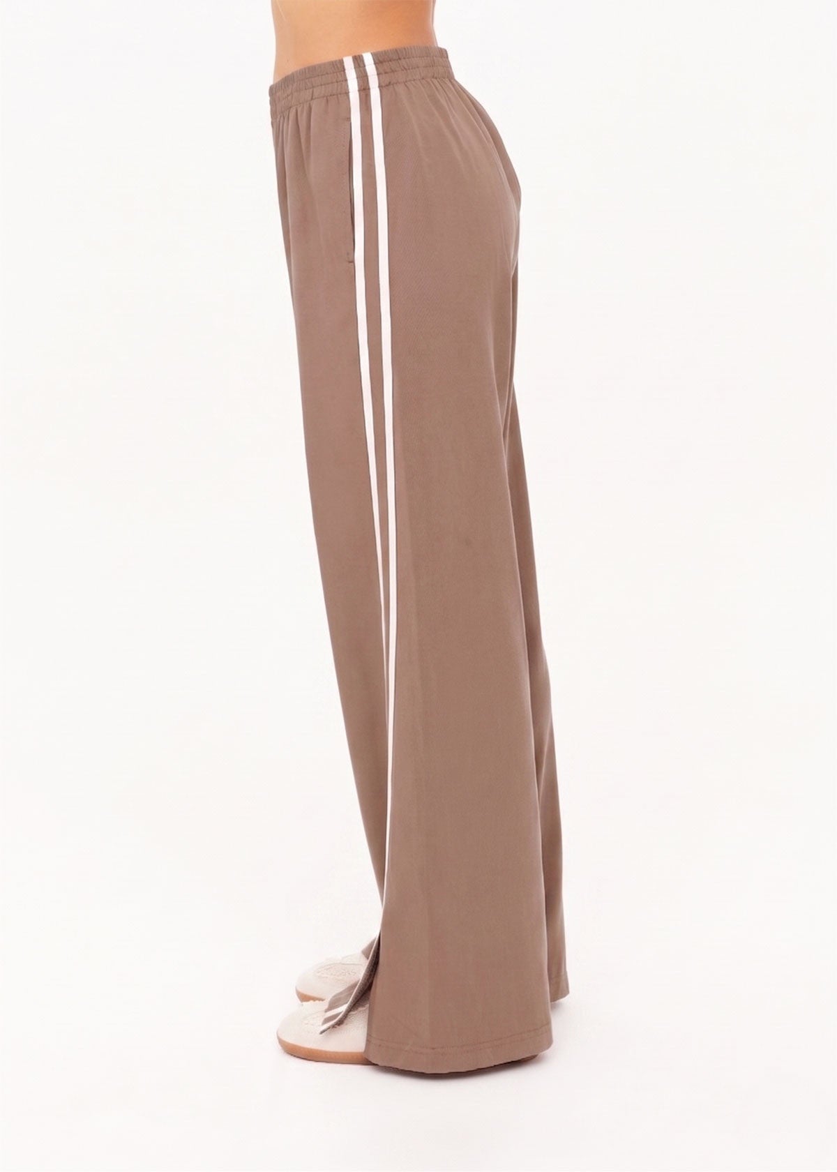 A person stands sideways against a plain white background, showcasing only their lower body and feet in The Upside JULIET PANT S26—loose, lightweight twill pants with an elastic waist and two white side stripes.