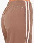 Close-up of a person wearing The Upside JULIET PANT S26 in brown twill with an elasticated waist, two white vertical side stripes, and a small white logo near the waistband. The background is plain and light-colored.