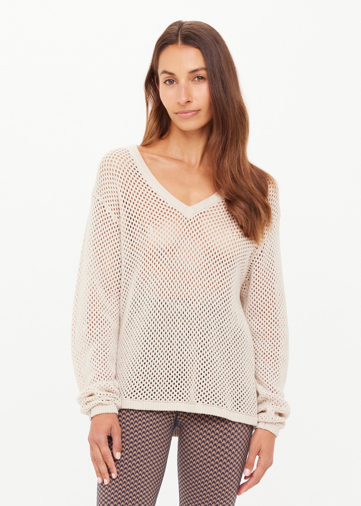 A woman with long brown hair wears The Upside's JOSEPHINE V NECK KNIT—a cream, open-knit relaxed sweater—paired with patterned leggings, standing against a white background and gazing neutrally at the camera.