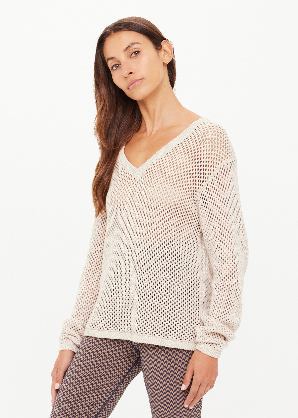 A woman with long brown hair and a neutral expression poses against a white background wearing The Upside's JOSEPHINE V NECK KNIT, a cream-colored, lightweight open knit sweater, layered over a white top and patterned pants.