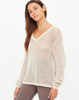 A woman with long brown hair and a neutral expression poses against a white background wearing The Upside's JOSEPHINE V NECK KNIT, a cream-colored, lightweight open knit sweater, layered over a white top and patterned pants.