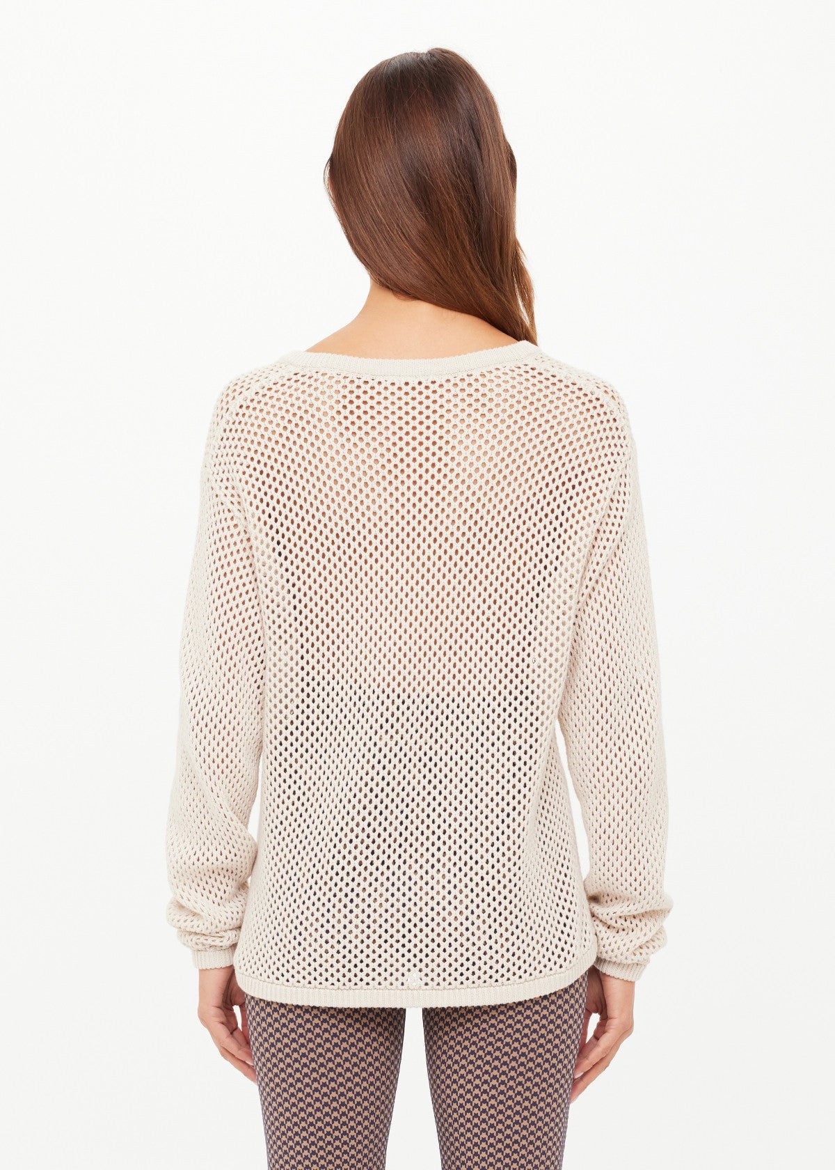 A woman with long brown hair faces away, wearing The Upside's JOSEPHINE V NECK KNIT—a loose, cream open-knit sweater with a relaxed V neckline—paired with patterned brown leggings against a plain white background.