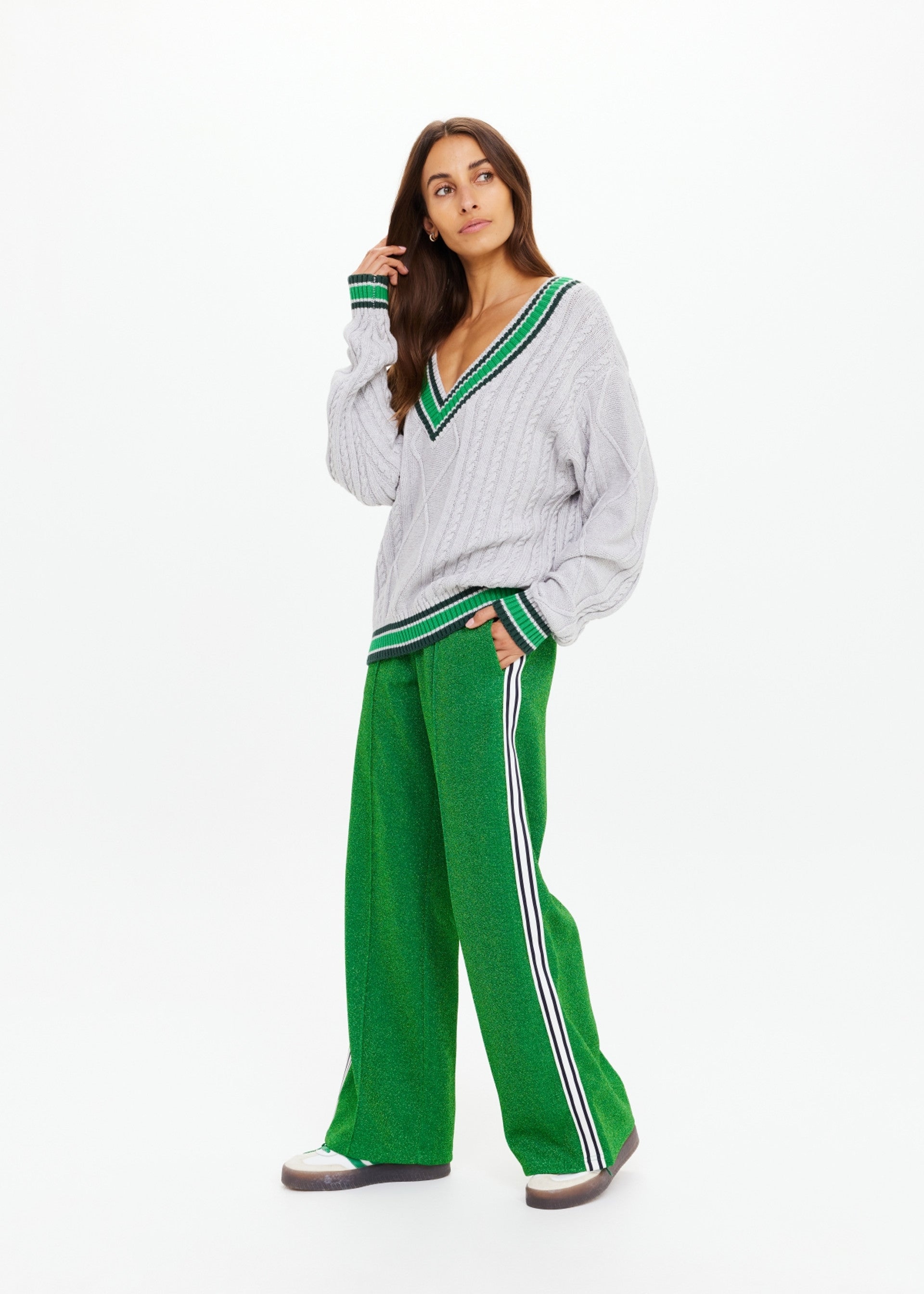 A woman stands against a white background wearing The Upside's wide-leg GALAXIA LORETTA PANT in green with white and navy side stripes, paired with a loose gray cable-knit sweater featuring Amazon Green trim. She looks to the side, hand near her chin.