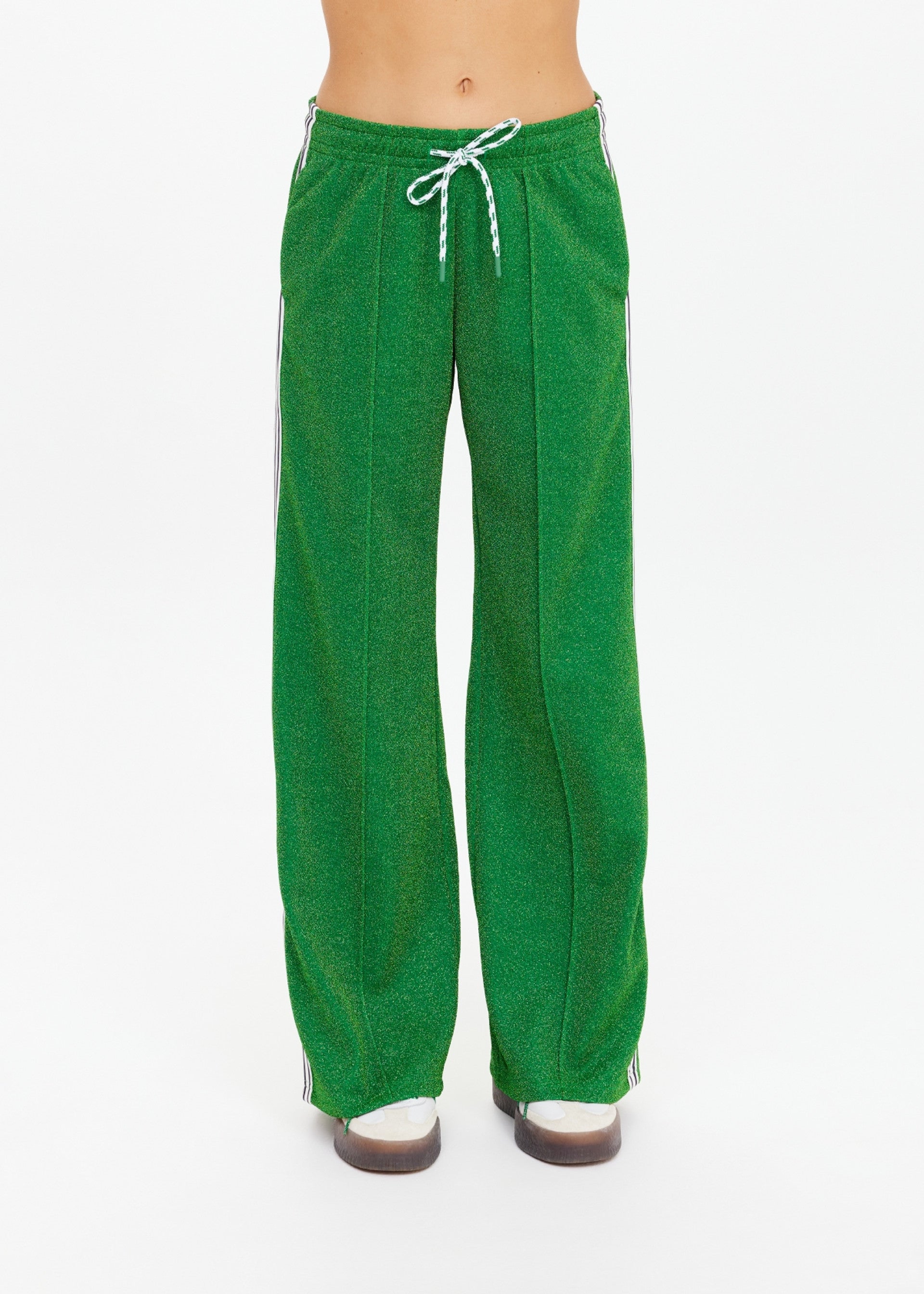 A person wearing The Upside GALAXIA LORETTA PANT in Amazon Green, featuring a drawstring waist and white side stripes, stands against a white background with only the lower torso and legs visible.