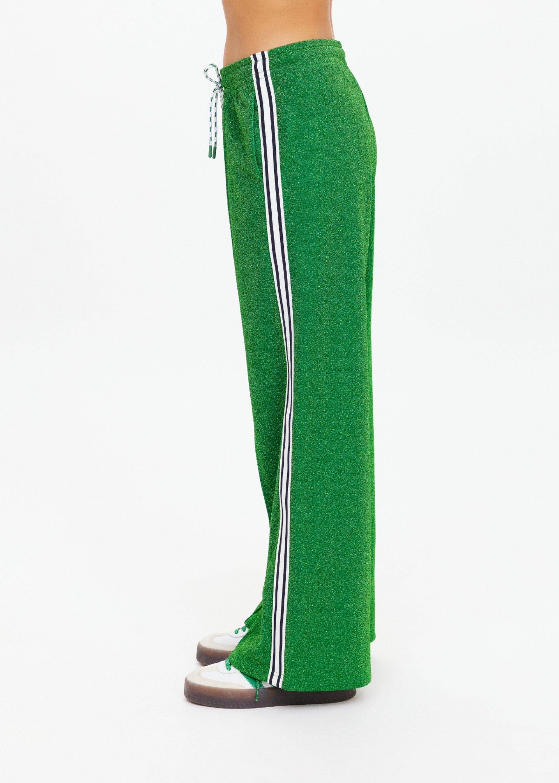 Person wearing The Upside GALAXIA LORETTA PANT in Amazon Green with white and black side stripe, paired with white socks and brown shoes, standing sideways against a plain white background.