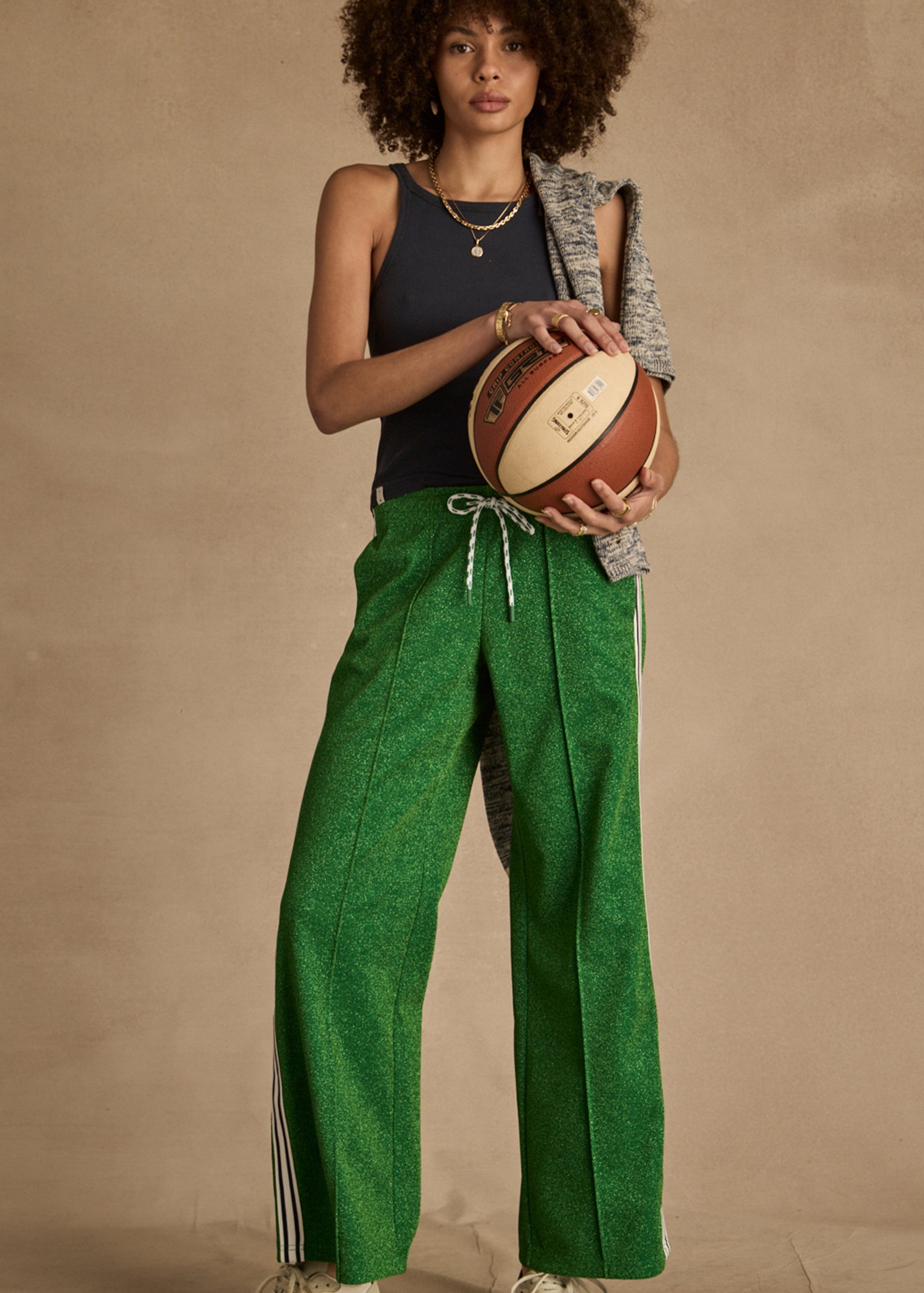Wearing a black tank top and The Upside's GALAXIA LORETTA PANT in Amazon Green, a person with curly hair stands against a neutral background, holding a basketball with a gray sweater draped over their shoulder.
