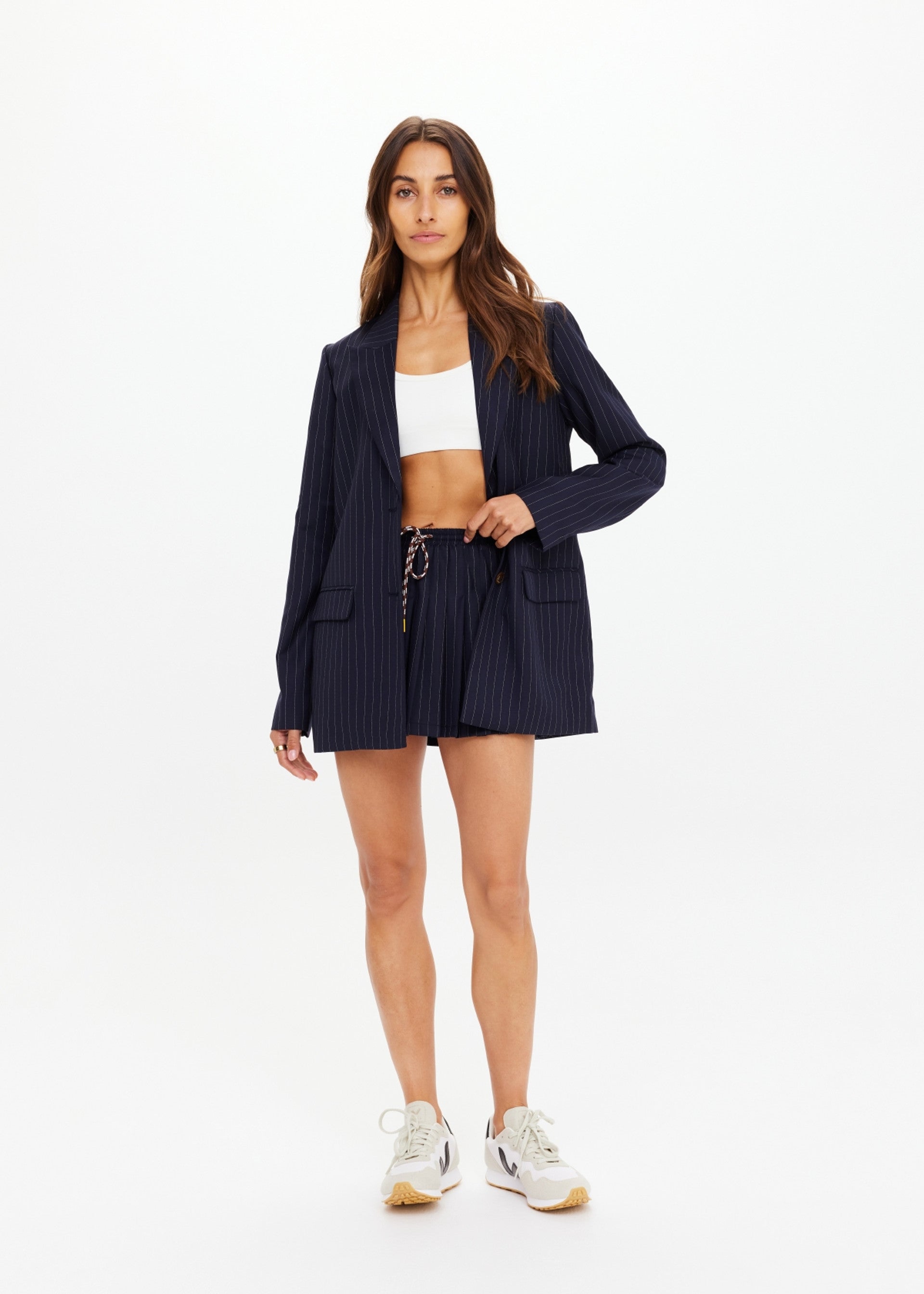 A woman models The Upside's WOOL BOYFRIEND BLAZER in navy pinstripe with matching shorts, paired with a white crop top and sneakers, highlighting its year-round versatility.