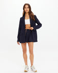 A woman models The Upside's WOOL BOYFRIEND BLAZER in navy pinstripe with matching shorts, paired with a white crop top and sneakers, highlighting its year-round versatility.