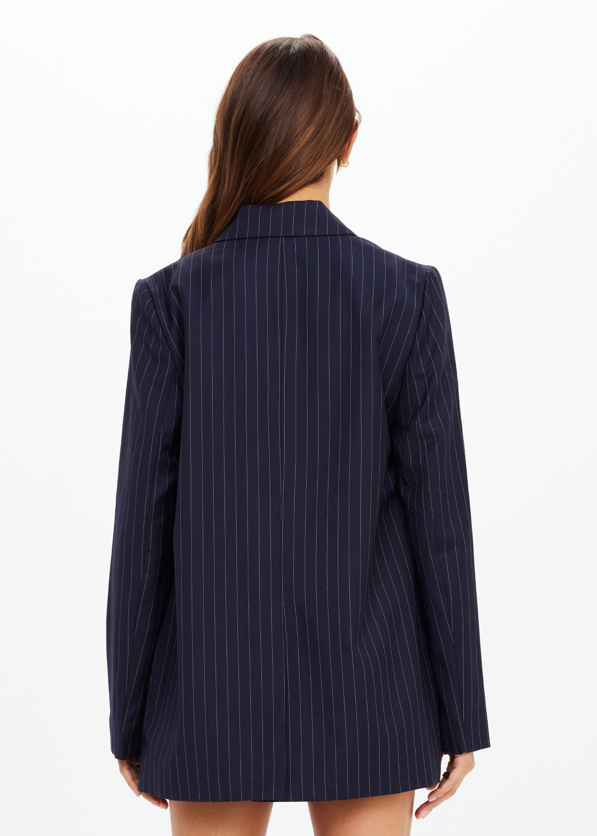 A woman with long brown hair stands facing away, wearing The Upside's WOOL BOYFRIEND BLAZER in dark navy. This Merino wool blazer offers year-round versatility against a plain white background. No visible pants.