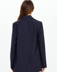 A woman with long brown hair stands facing away, wearing The Upside's WOOL BOYFRIEND BLAZER in dark navy. This Merino wool blazer offers year-round versatility against a plain white background. No visible pants.