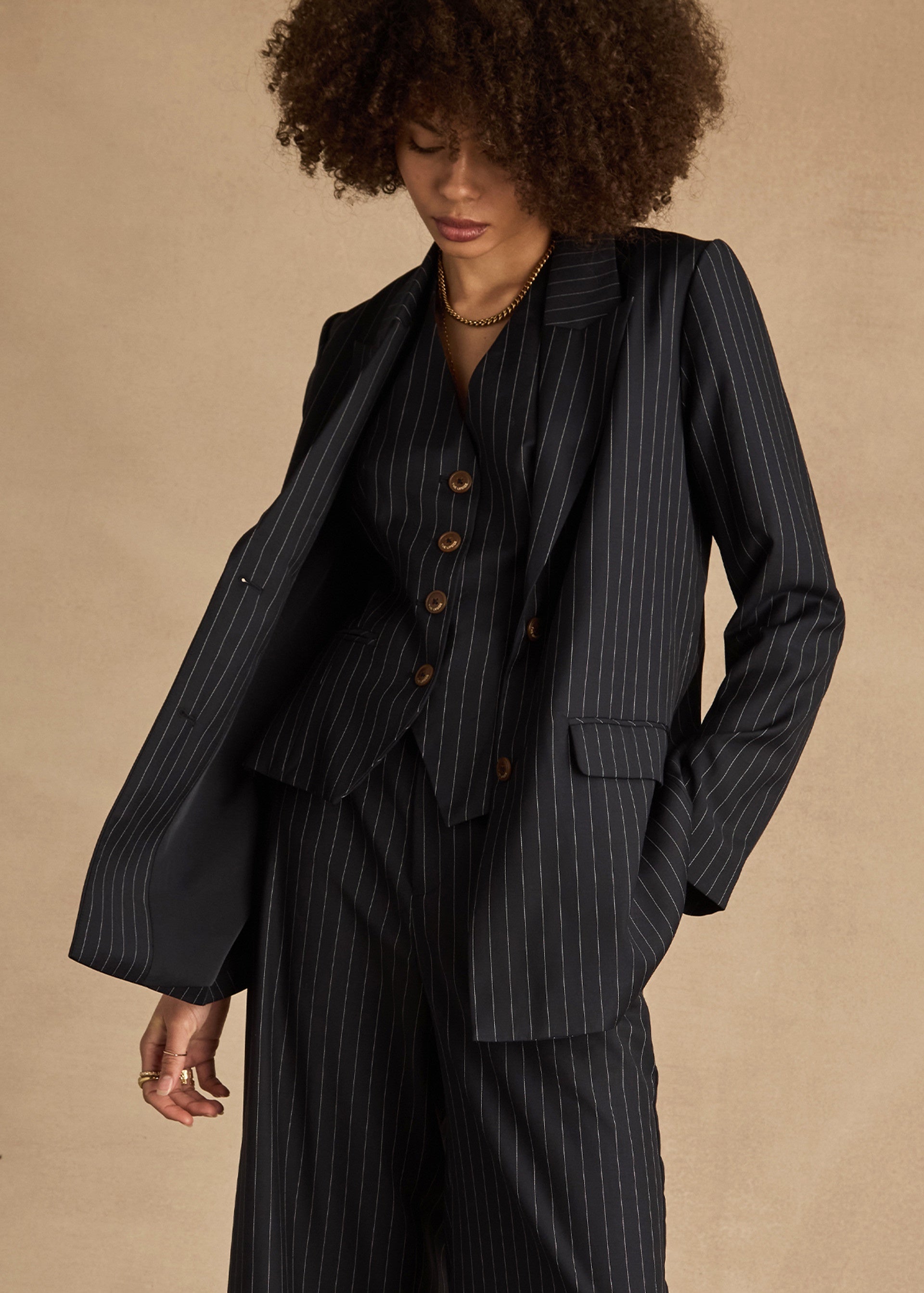 A person with curly hair wears The Upside’s WOOL BOYFRIEND BLAZER, paired with wide-leg pants and a matching vest, standing against a beige background and looking down.