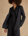 A person with curly hair wears The Upside’s WOOL BOYFRIEND BLAZER, paired with wide-leg pants and a matching vest, standing against a beige background and looking down.