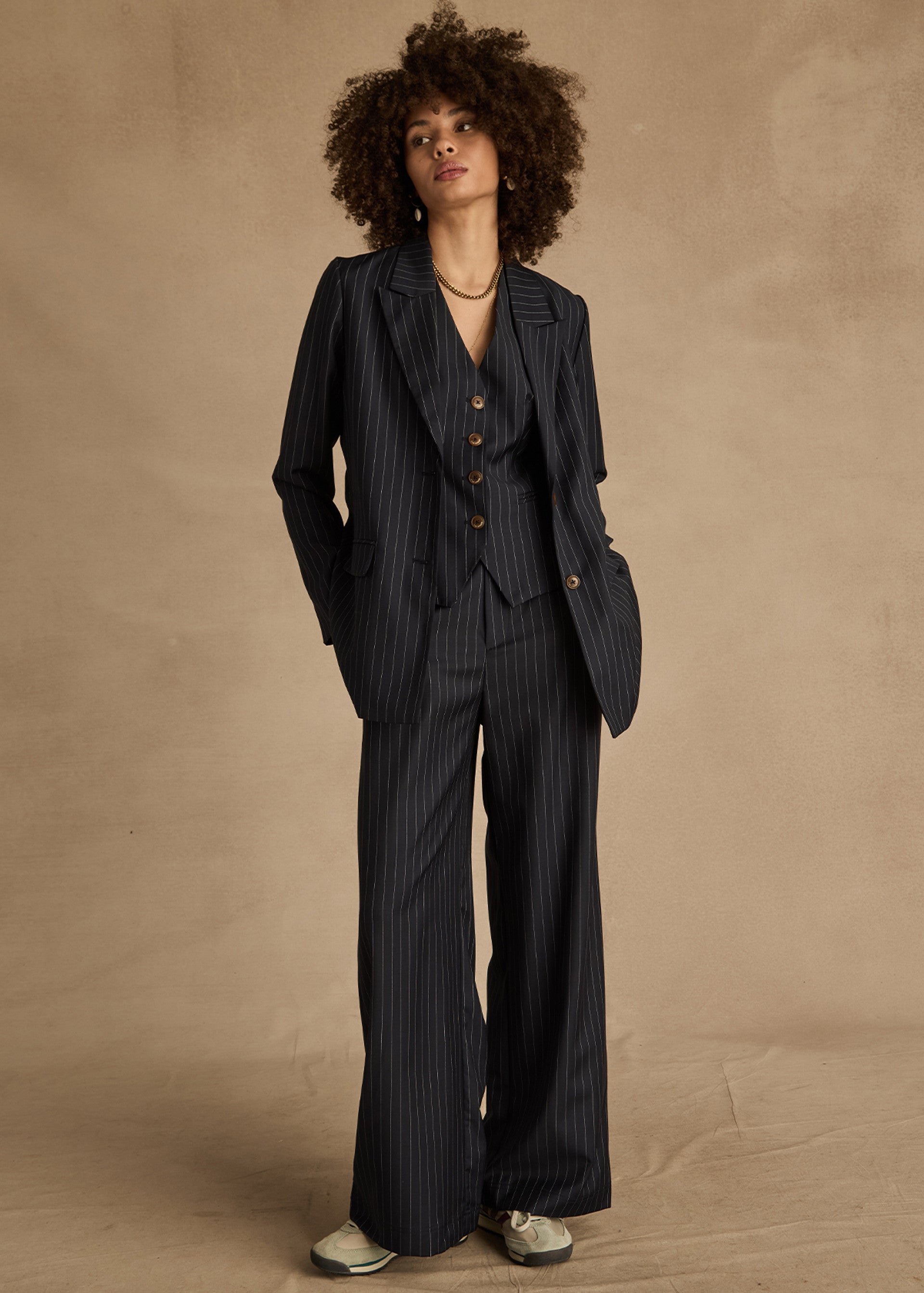A person with curly hair wears The Upside's WOOL BOYFRIEND BLAZER—a dark pinstripe, wrinkle-resistant suit. With hands in their pockets and sneakers on, they confidently stand against a neutral beige backdrop.