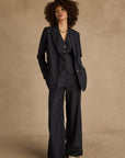 A person with curly hair wears The Upside's WOOL BOYFRIEND BLAZER—a dark pinstripe, wrinkle-resistant suit. With hands in their pockets and sneakers on, they confidently stand against a neutral beige backdrop.