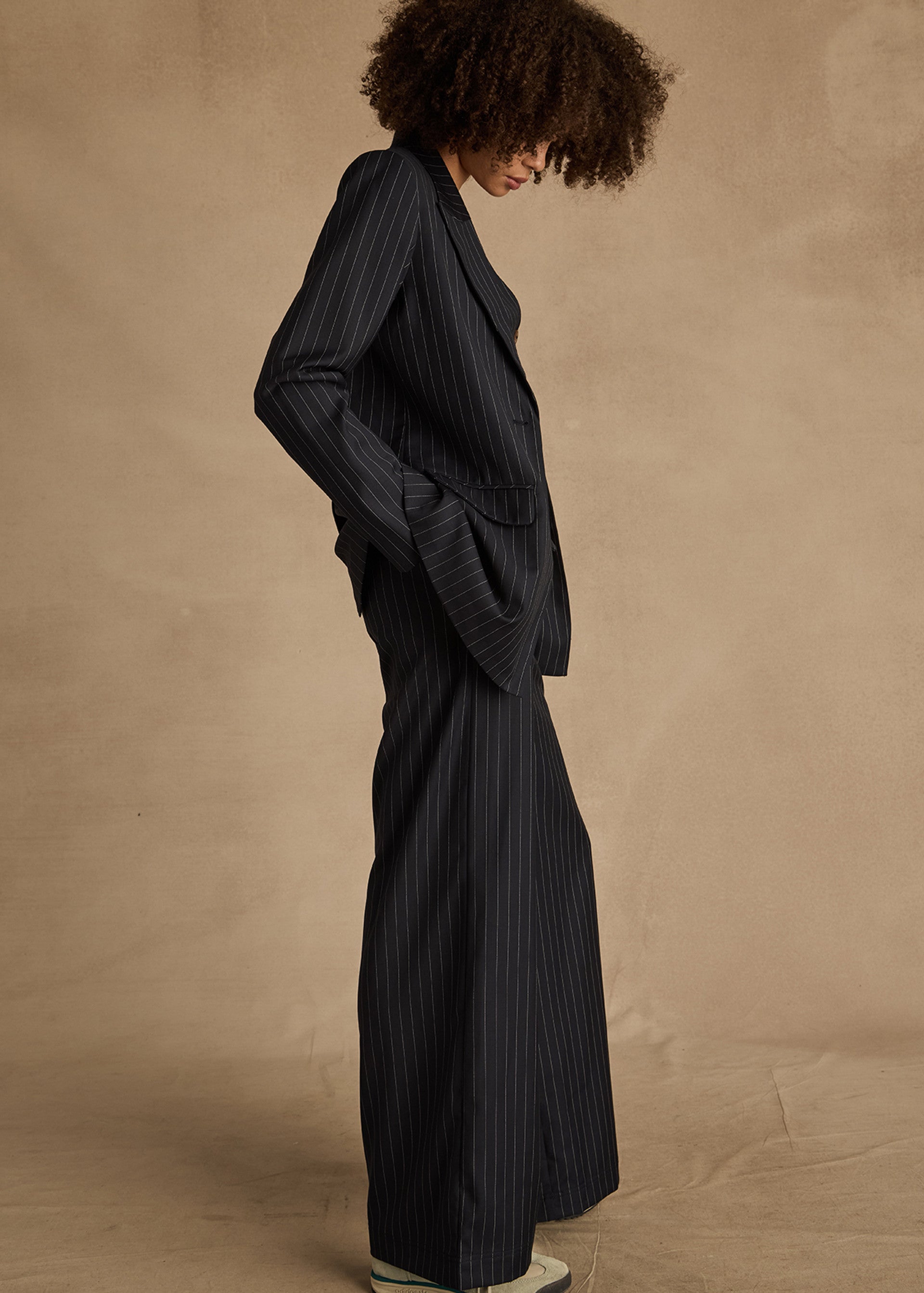 A person with curly hair wears The Upside's WOOL BOYFRIEND BLAZER in dark pinstripe and light shoes, standing in profile against a beige background—an ideal choice for year-round wearability.