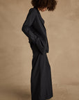 A person with curly hair wears The Upside's WOOL BOYFRIEND BLAZER in dark pinstripe and light shoes, standing in profile against a beige background—an ideal choice for year-round wearability.