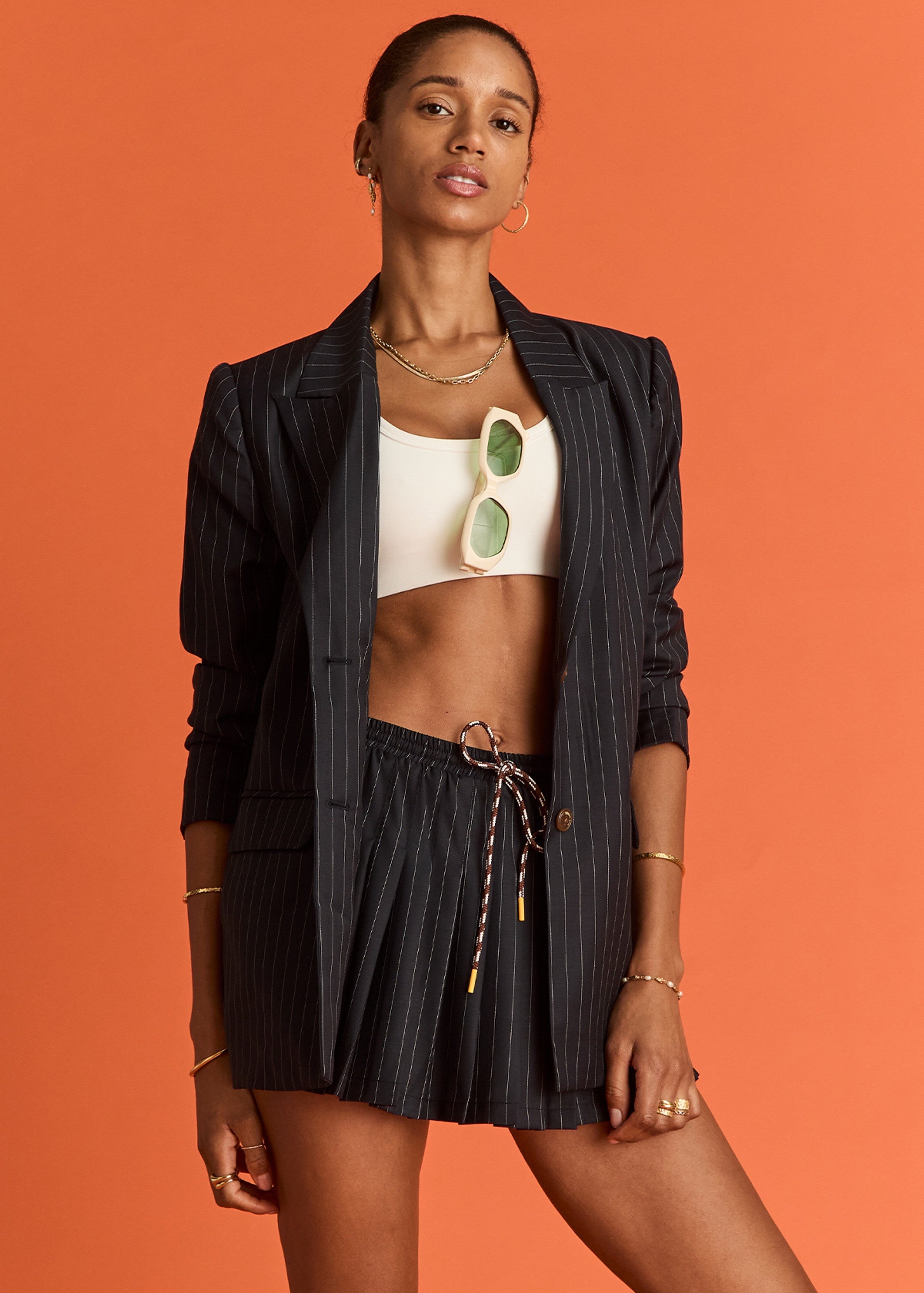 A woman poses against an orange background, wearing The Upside's WOOL CADIZ PLEATED SKIRT with a black pinstripe blazer, white crop top, sunglasses tucked into her top, and gold jewelry.