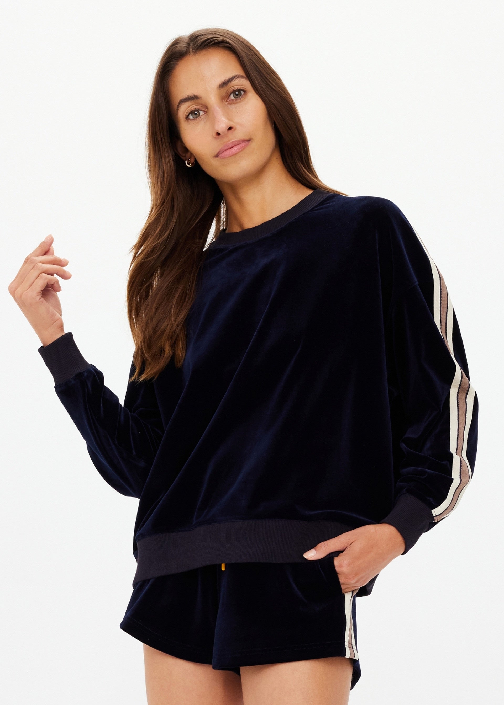 A woman wears The Upside MONACO LISEL CREW NECK SWEATER in navy velour with striped sleeves and matching recycled polyester shorts, standing against a white background and looking at the camera with a relaxed expression.