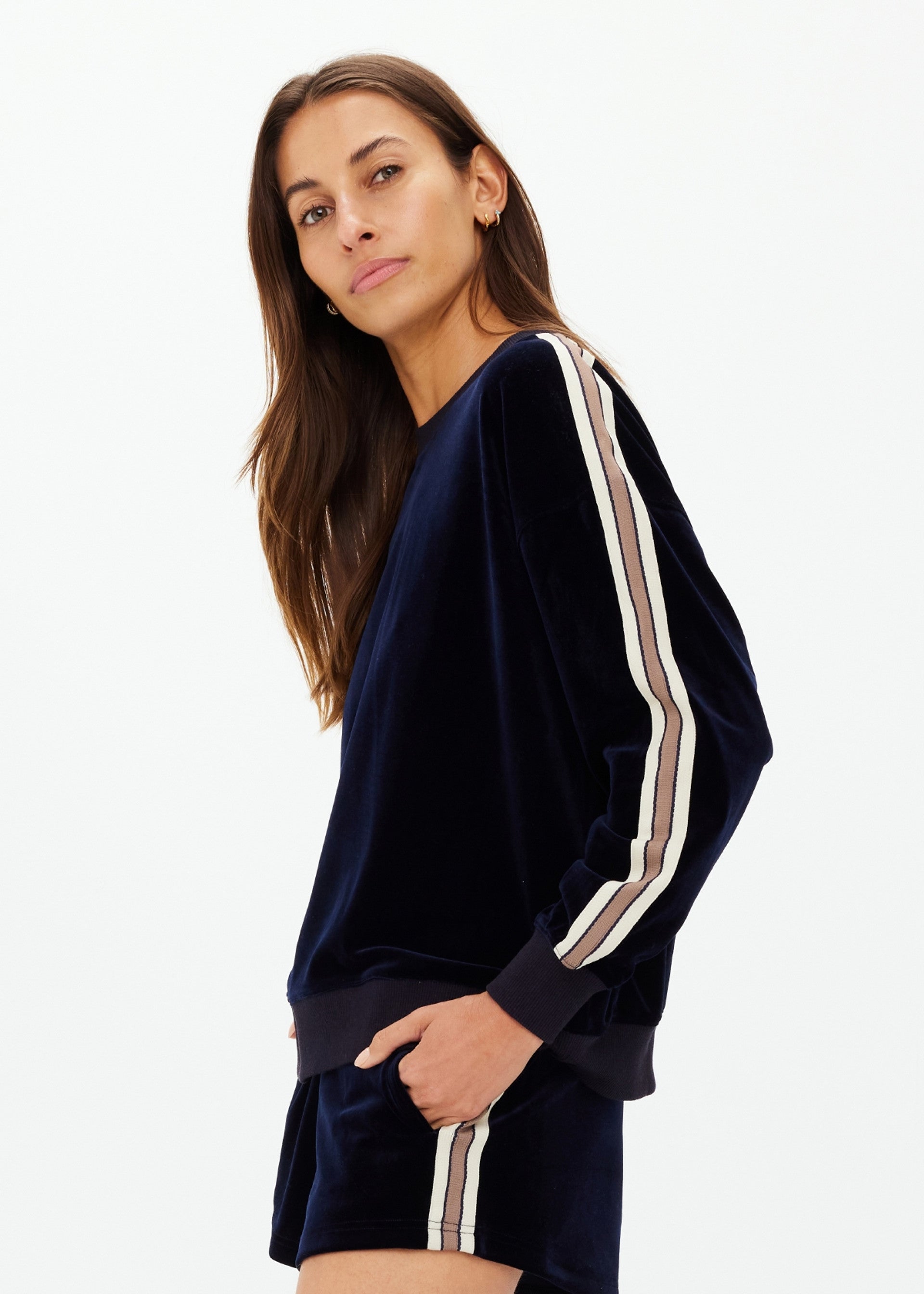 A woman with long brown hair stands sideways in The Upside MONACO LISEL CREW NECK SWEATER, a navy velour sweatshirt made from recycled polyester with beige and white striped sleeves, paired with matching shorts. She looks slightly toward the camera.