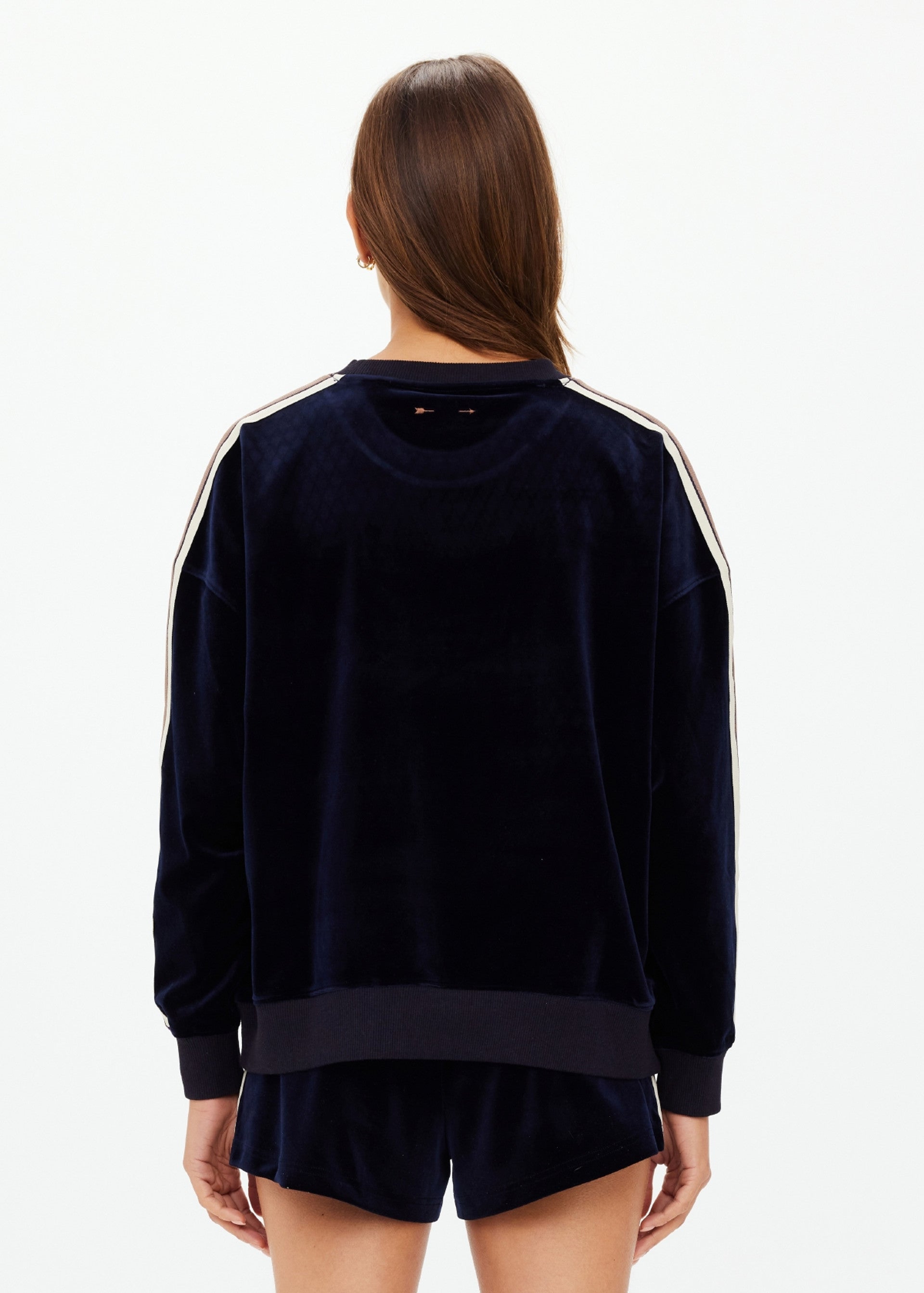 A woman with long brown hair, seen from behind, is wearing The Upside’s MONACO LISEL CREW NECK SWEATER in navy velour with white sleeve stripes and matching recycled polyester shorts, set against a plain white background.