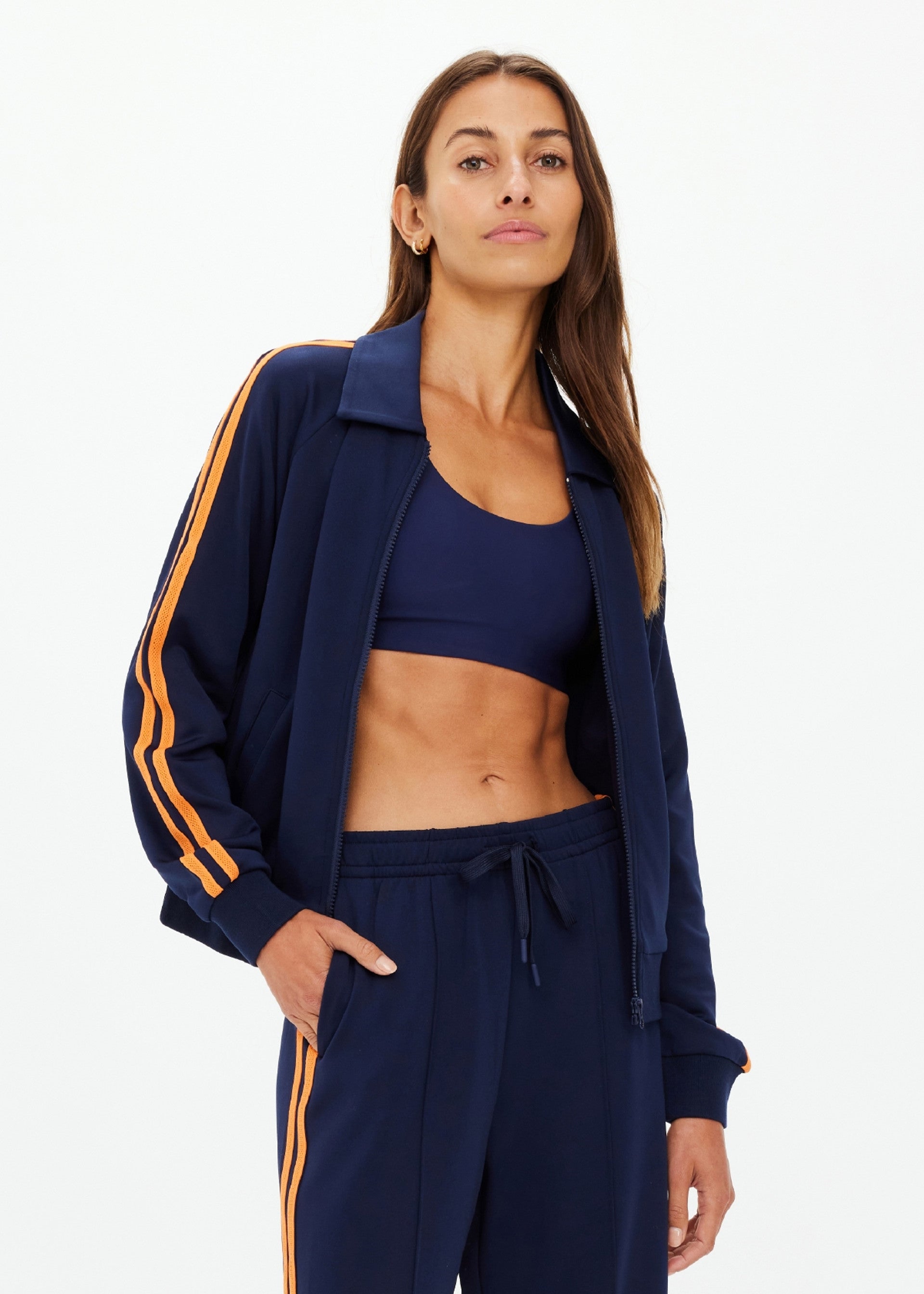 A woman stands confidently against a white background wearing The Upside's VITESSE PARI ZIP JACKET—a navy sports jacket with matching pants, featuring orange stripes in slinky fabric.