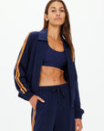 A woman stands confidently against a white background wearing The Upside's VITESSE PARI ZIP JACKET—a navy sports jacket with matching pants, featuring orange stripes in slinky fabric.