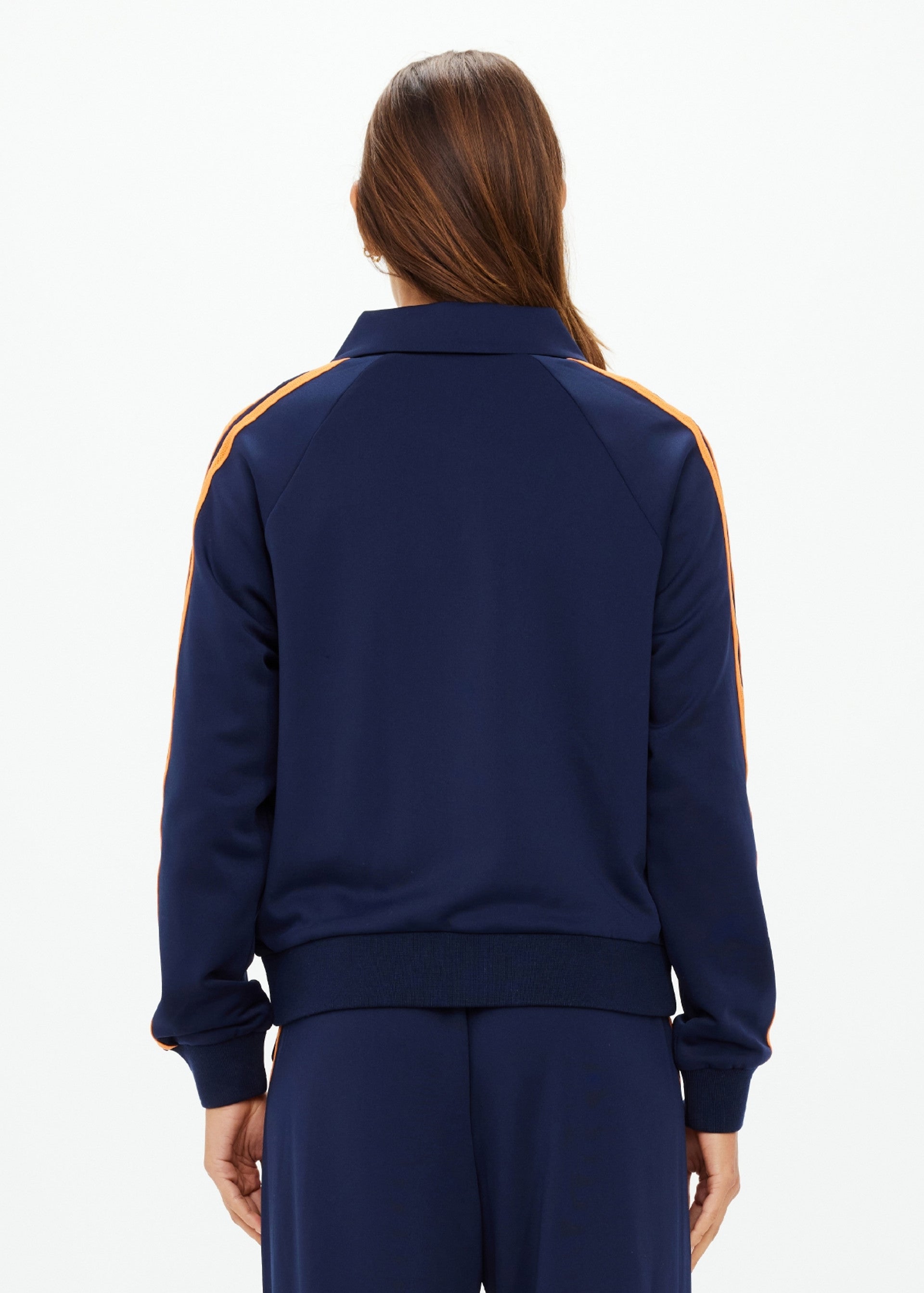 A person with long brown hair, seen from behind, wears The Upside's VITESSE PARI ZIP JACKET in navy with orange sleeve stripes and matching slinky pants, set against a plain white background.